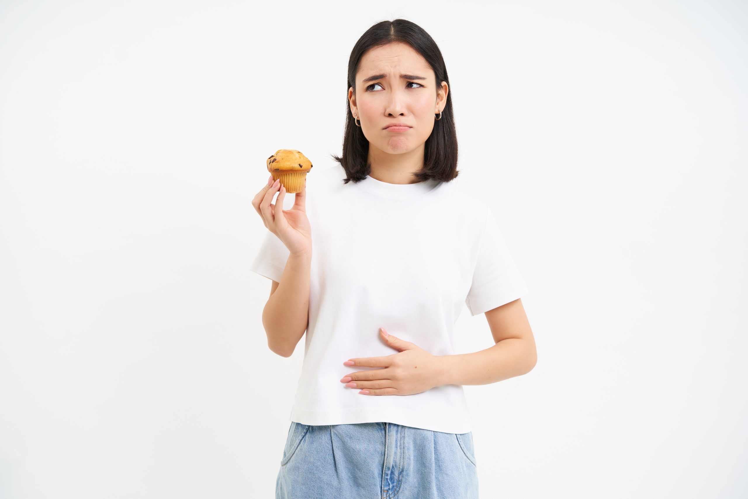 Portrait of asian woman feeling pain in stomach, discomfort after food, holding cupcake and looking unhappy, white background.