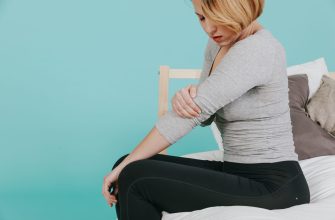 Woman seated on a bed, wearing a gray long-sleeve top and black leggings, resting her shoulder in pain against a teal wall.