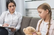 Therapist sitting with a clipboard talking to a girl on a couch, holding a blue pen for notes