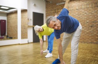 Two seniors perform a standing forward bend stretch in a gym studio with a wooden floor and brick wall background, each reaching toward the floor.