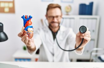 Healthcare professional holds a colorful human heart model, using a stethoscope in the background for a medical demonstration.