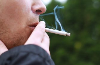 Close-up of a person smoking a cigarette outdoors, with smoke curling from the lit end against a green blurred background.”