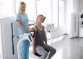 Healthcare worker in blue scrubs places a blood pressure cuff on a seated patient during a medical test or rehab session