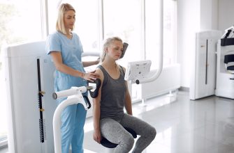Healthcare worker in blue scrubs places a blood pressure cuff on a seated patient during a medical test or rehab session