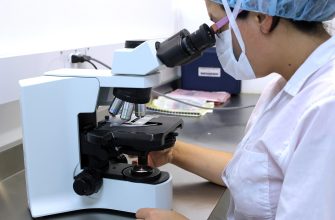 Scientist in a lab coat wearing a mask and hair cover looks into a light blue compound microscope in a laboratory setting.