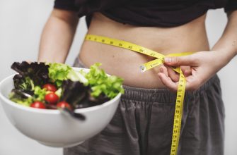Person uses a yellow measuring tape to measure their waist while holding a bowl of salad, emphasizing health and dieting goals.