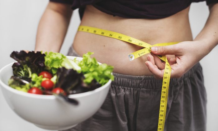 Person uses a yellow measuring tape to measure their waist while holding a bowl of salad, emphasizing health and dieting goals.