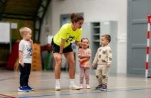 Coach in a bright neon shirt crouches to speak with four young children in a gymnasium during indoor activity.