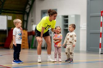 Coach in a bright neon shirt crouches to speak with four young children in a gymnasium during indoor activity.