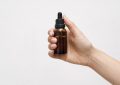 Hand holding a small amber glass dropper bottle with a black cap against a light background.
