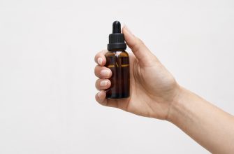 Hand holding a small amber glass dropper bottle with a black cap against a light background.