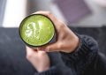 Hand holding a mug of frothy green matcha latte with a white foam leaf design on top.