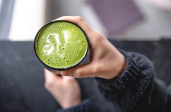 Hand holding a mug of frothy green matcha latte with a white foam leaf design on top.
