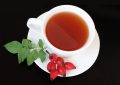 Top-down view of a white teacup with reddish tea on a matching saucer, accompanied by rose hip berries and green leaves on a black background.