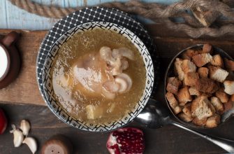 Bowl of clear soup with dumplings on a patterned plate, beside a bowl of croutons on a rustic wooden table.