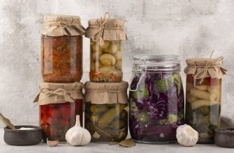 Assorted jars of homemade pickles and preserved vegetables with parchment lids tied in twine, arranged on a rustic surface.