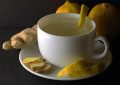 White teacup on a saucer with a lemon-peel garnish, surrounded by ginger and lemon against a dark background.
