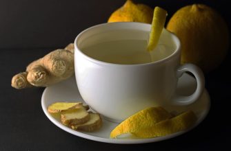 White teacup on a saucer with a lemon-peel garnish, surrounded by ginger and lemon against a dark background.