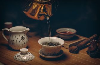 Teapot pours hot water into a white teacup on a wooden table, with tea leaves in a bowl nearby.