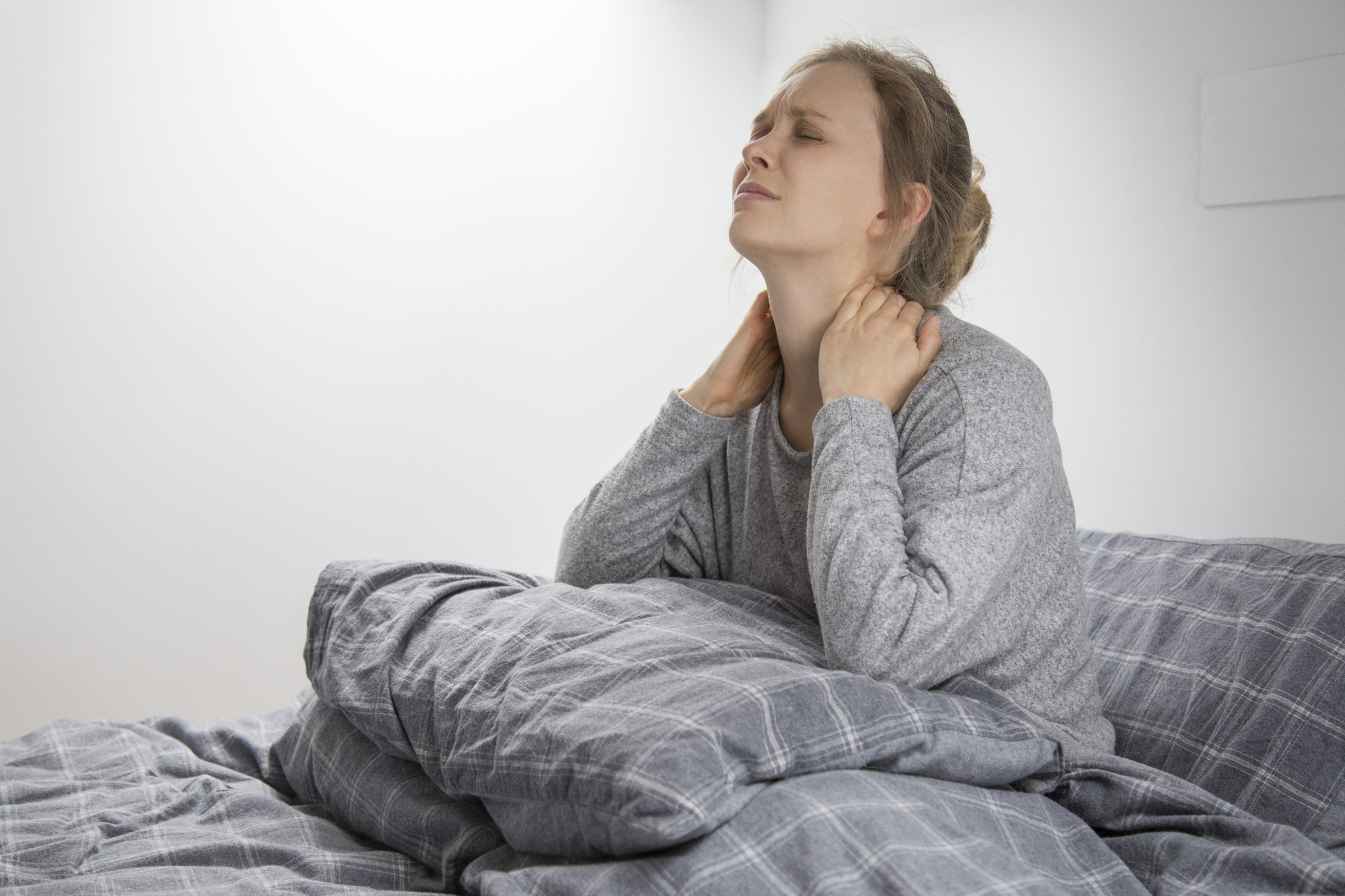 Tired sick young Caucasian woman in grey homewear sitting on bed with closed eyes, touching her neck, suffering from pain. Illness, pain concept